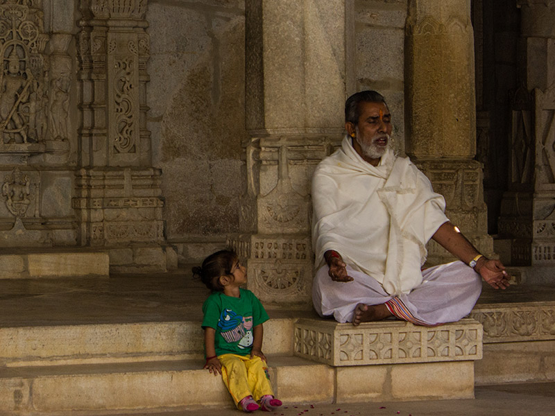 There's an old man with a white beard with his eyes closed. He seems to be praying. A little girl is sitting next to her watching him.