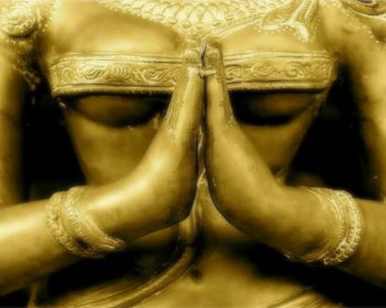 Close-up photo of the torso of a beautiful antique Indian statue with hands in the praying, or Namaste, gesture. The statue is golden appears to represent a female person or deity..