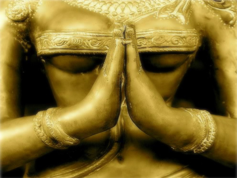 Close-up photo of the torso of a beautiful antique Indian statue with hands in the praying, or Namaste, gesture. The statue is golden appears to represent a female person or deity..
