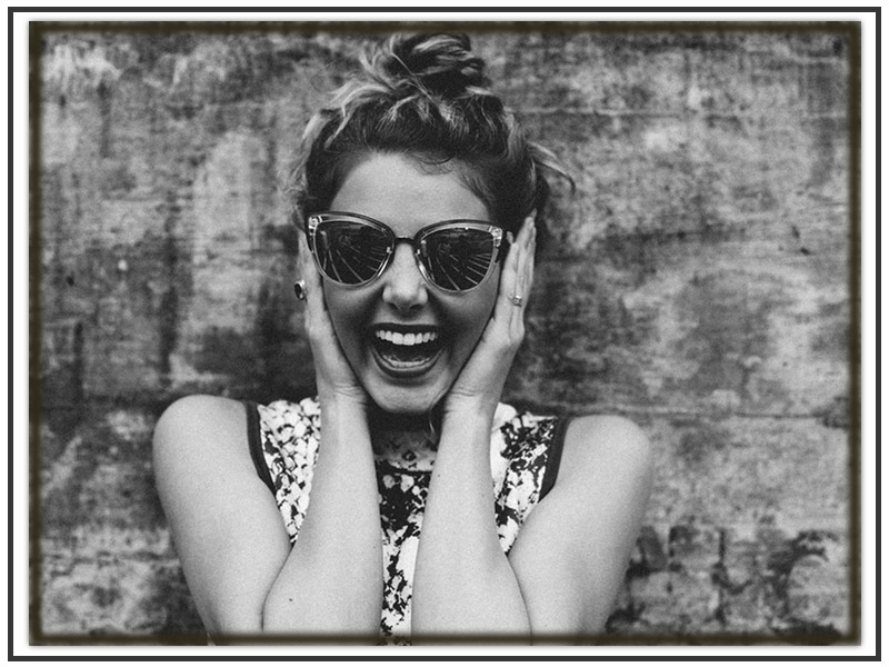 A portrait of a young woman smiling with her mouth open broadly. She is wearing glasses. Her hands are on her face. The image is in black and white