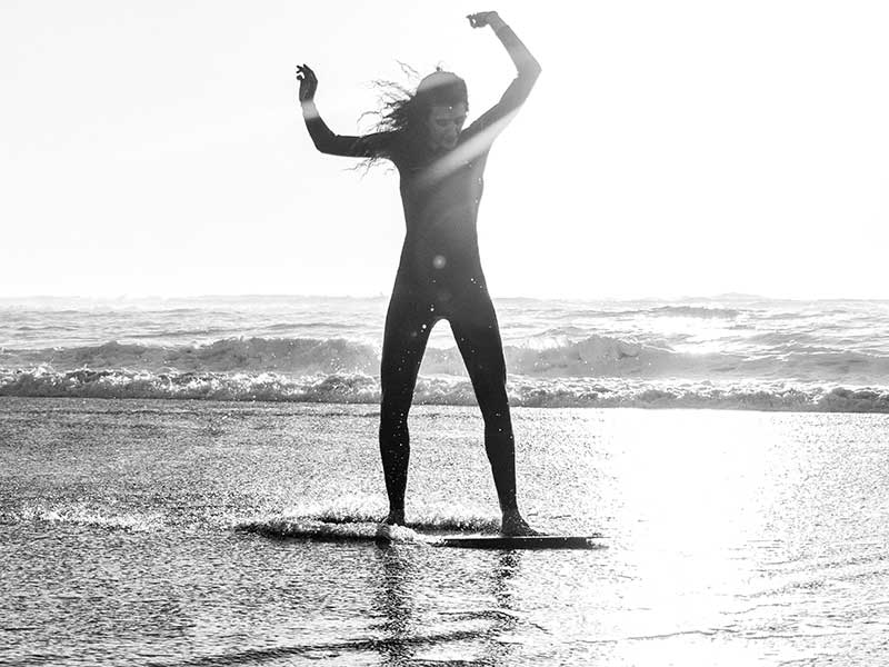 A sea surfer on a skimboard in the middle of the sea seems to be surfing alone. The sea surfer seems to be a young woman but the photo is in black and white making it difficult to tell.