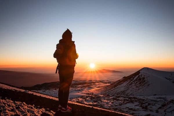 A person wearing a backpack and knit cap stands above a vista of mountain tops, looking towards the rising sun. The sky is blue, white and orange.
