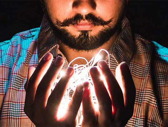 A bearded man bolds light fibers in his cupped hands. The light illuminates his face from the nose down.