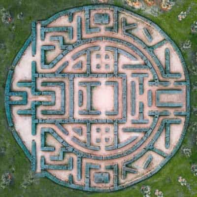 Aerial shot of an intriguing circular shrub maze surrounded by greenery. The floor of the maze is a light tan color.