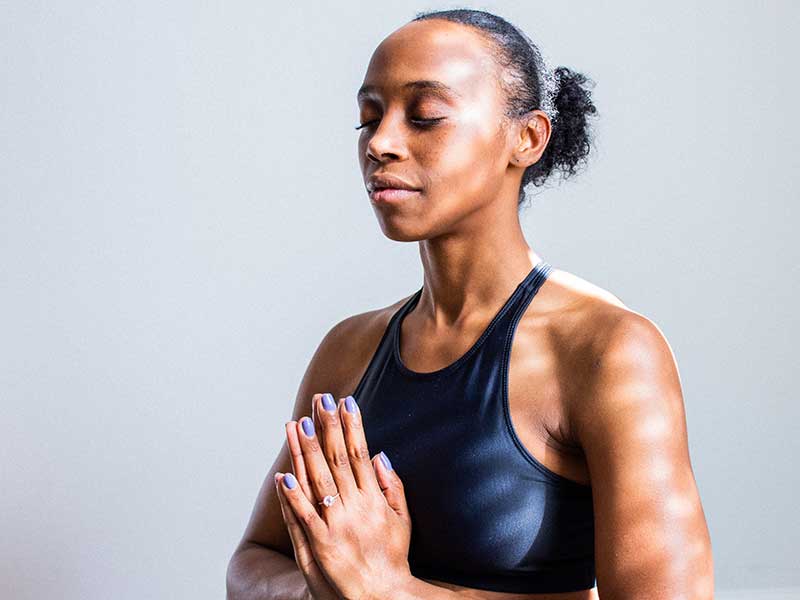 A young black woman wearing a dark colour sport bra. Her eyes are closed and the palm of her hands are in contact. She seems to be meditating.