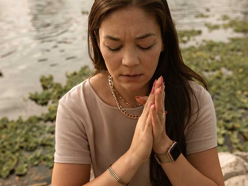 A young woman with long brown hair is seated outdoors at a peaceful riverbank, her hands folded as if to meditate, but her brow is furrowed.