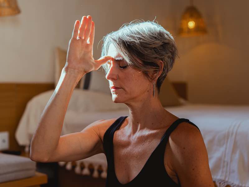 A person with short hair and dangling earrings sits with eyes closed, thumb to forehead, fingers point upwards.The background appears to be a bedroom with white walls and soft sconce lighting.