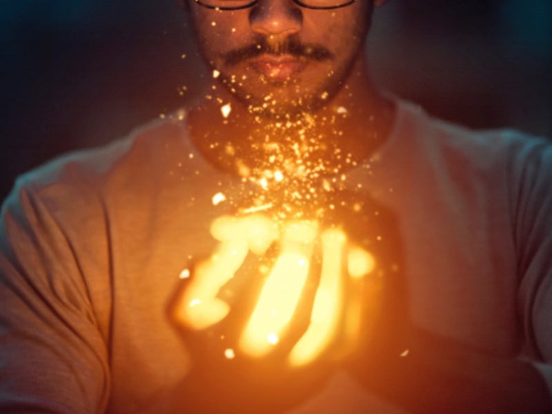 A close-up of a pair of hands appearing to hold fire sparks that glow bright orange and yellow. The half-face seen above the hands appears to be male and is calm and relaxed.