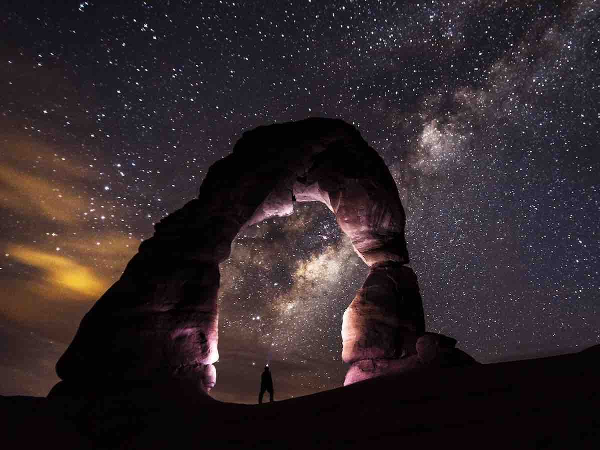 A person is standing under a huge natural rock arch formation at night. The inner sides and top of the arch are light purple. In the background is an expansive vista of a star-filled sky.