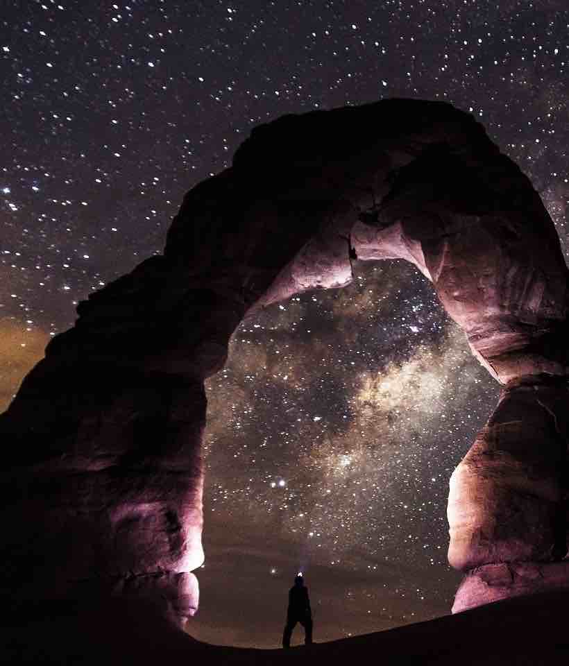 A person is standing under a huge natural rock arch formation at night. The inner sides and top of the arch are light purple. In the background is an expansive vista of a star-filled sky.