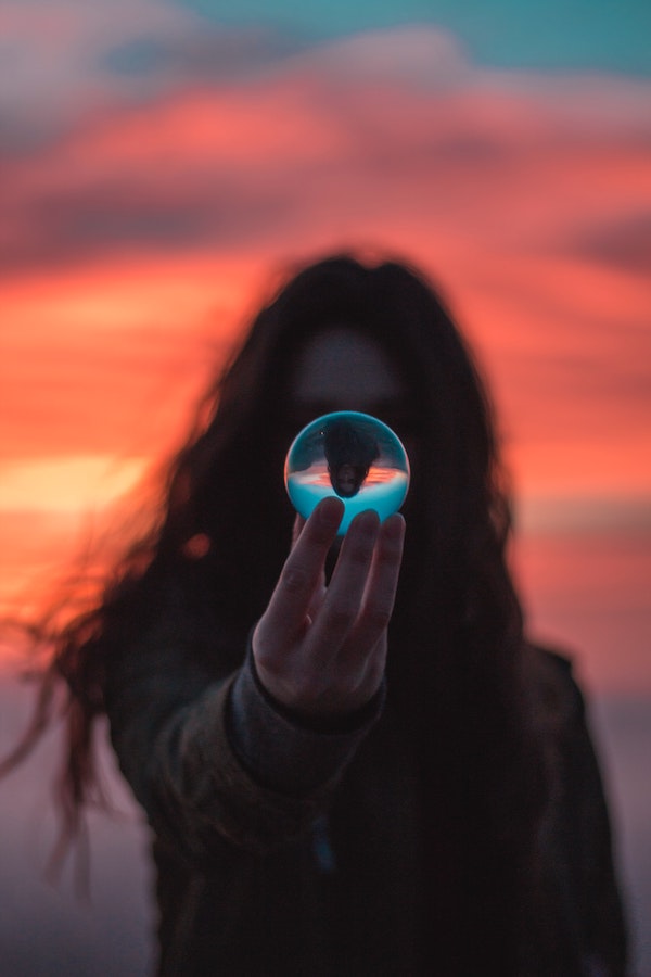 A person with wild long hair offers a sphere that blocks a clear view of their face. The sphere reflects an upside down image of sky and the person. The background is orange and pink as though during sunrise or sunset.