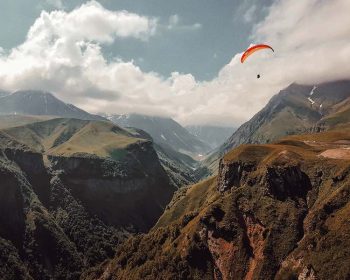 A dramatic cloud-filled sky with a single open red parachute gliding gracefully across the vast and peaceful canyon below.