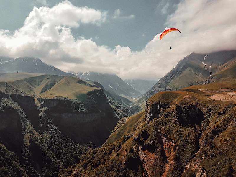 A dramatic cloud-filled sky with a single open red parachute gliding gracefully across the vast and peaceful canyon below.