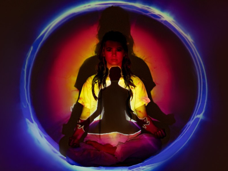 A person sits crossed legged inside a sphere of different color lights, seeming to be in a trance-like state