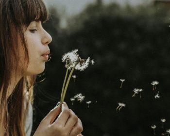 A young woman is in profile against a dark background, gently blowing dandelion seeds.