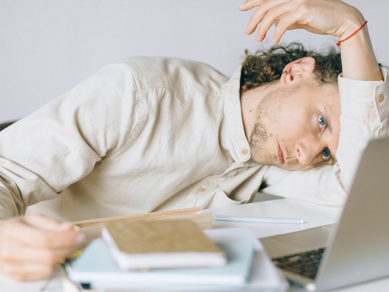 A young man represents decision fatigue by being slumped over his desk littered with books in a messy pile. He looks unhappy as he gazes with tired eyes at his laptop.